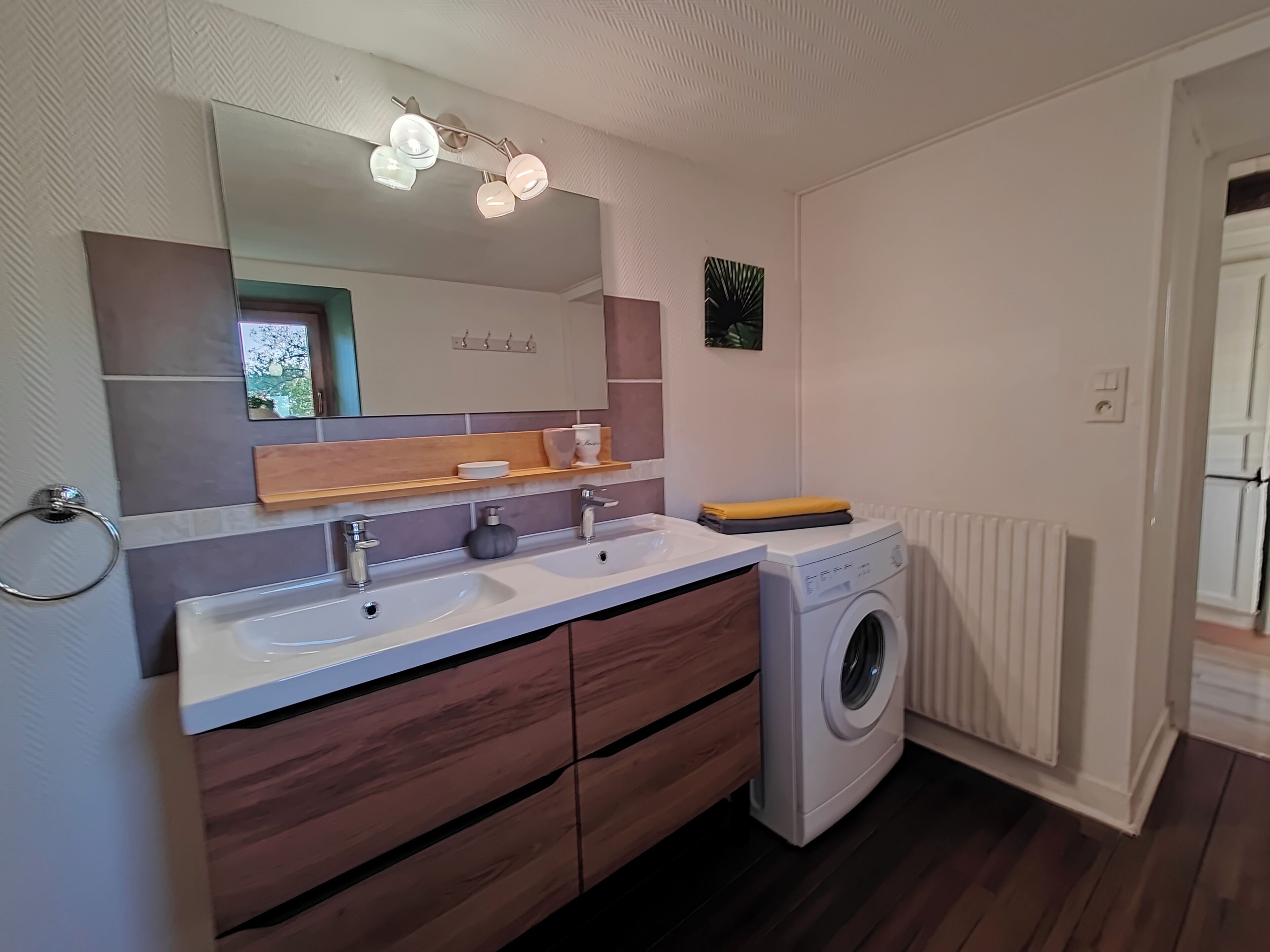 Un baño moderno con dos lavamanos, un espejo, un perchero para toallas, un estante pequeño con artículos de aseo y una lavadora.