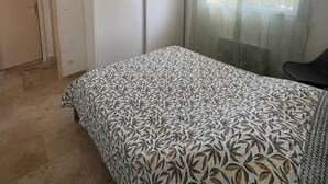 Un dormitorio con una colcha estampada, una lámpara colgante, una puerta y una ventana con cortinas translúcidas.
