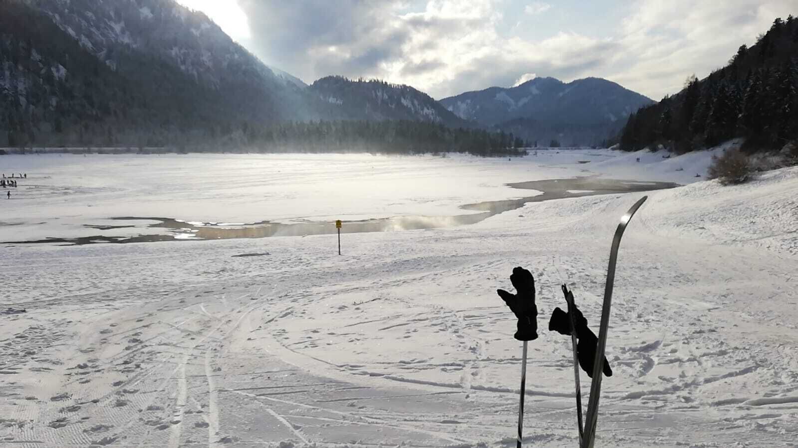 Schneebedeckte Landschaft mit einem zugefrorenen See, Bergen im Hintergrund und einem Paar Ski, die an einem Pfahl lehnen.