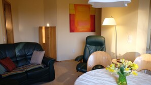 A living room with a black leather sofa, a green armchair, a wooden table with a vase of yellow flowers, and a floor lamp. - Apartment in a green idyll on the edge of Chemnitz (Chemnitz)