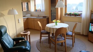 A cozy living room with a dining table, chairs, a black leather chair, a small side table, a lamp, a TV, and a window with curtains. - Apartment in a green idyll on the edge of Chemnitz (Chemnitz)