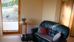 A living room with a black leather sofa, a small side table with a plant, and a view of a garden through a glass door. - Apartment in a green idyll on the edge of Chemnitz (Chemnitz)