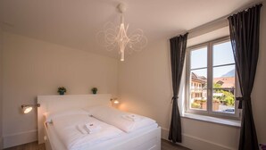 A bedroom with a bed, a window with curtains, a chandelier, and a view of buildings outside. - Spacious and central apartment with terrace (Matten bei Interlaken)