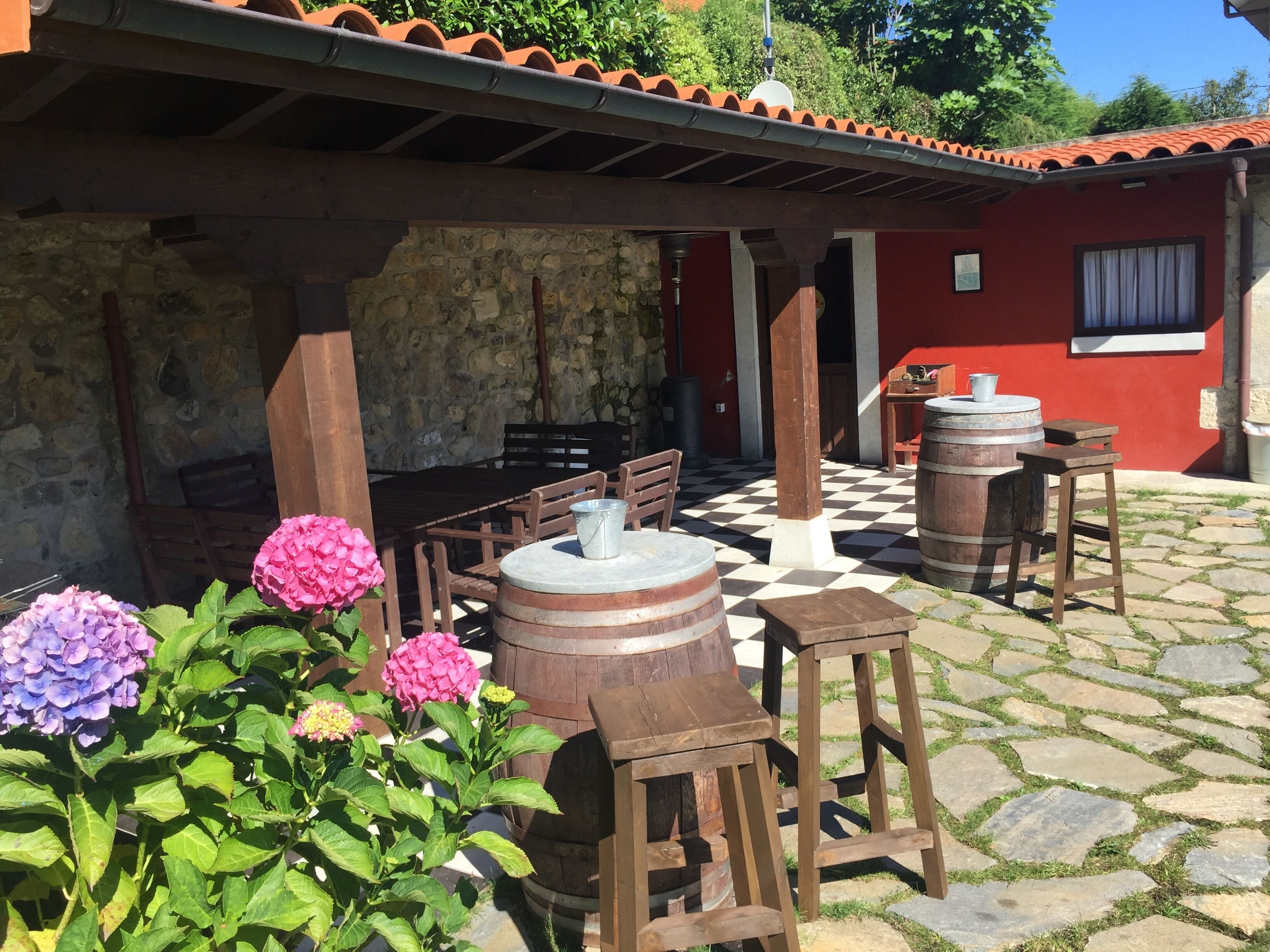 An outdoor patio with stone flooring, wooden furniture, and a red building in the background.
