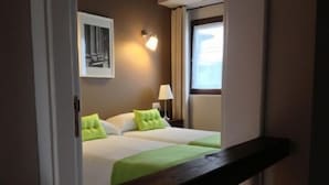 Habitación de hotel con dos camas, una mesita de noche con lámpara, un cuadro enmarcado en la pared y una ventana con cortinas.