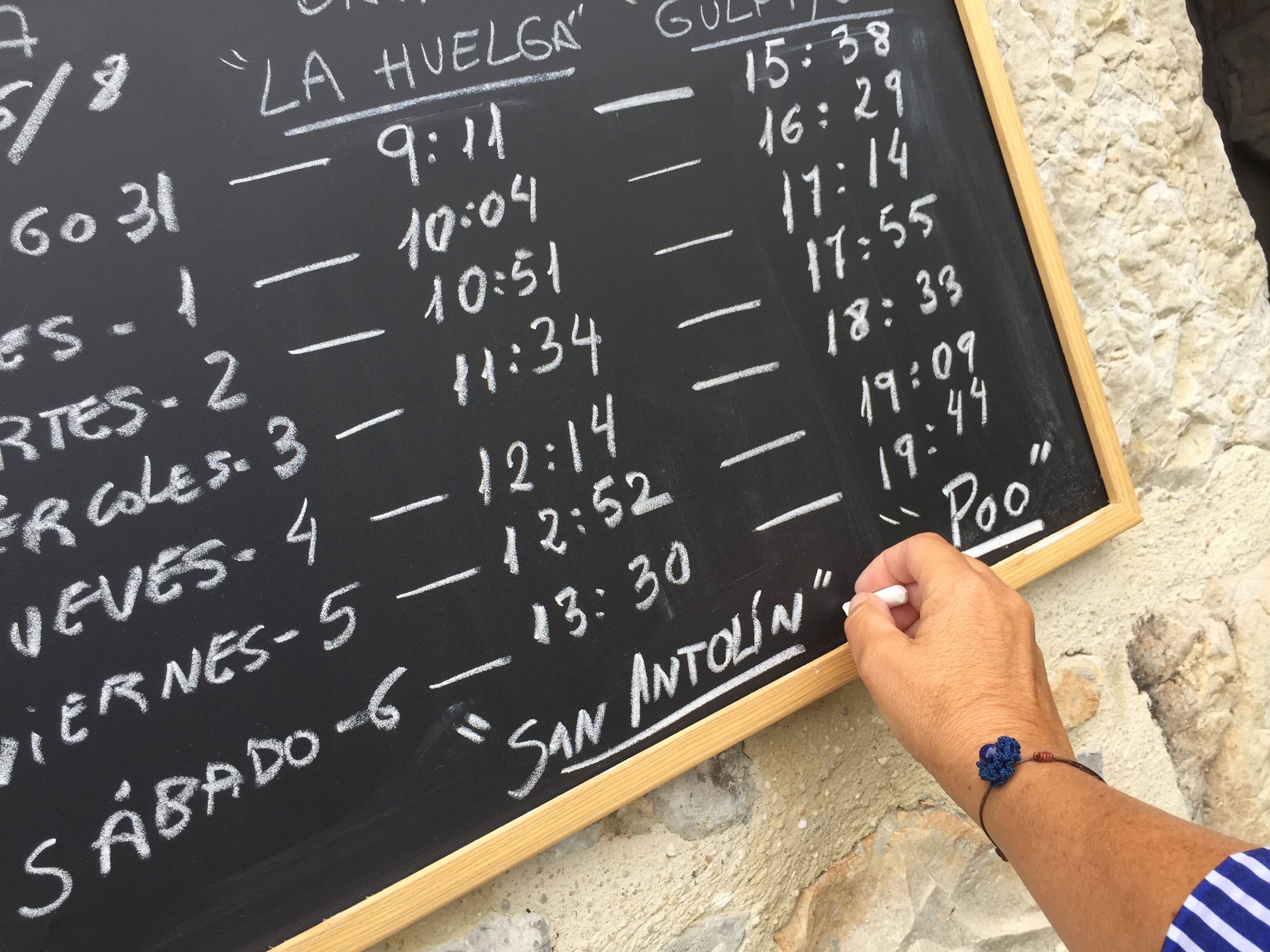 A blackboard with a schedule listing times and days, a hand holding it, and a stone wall background.