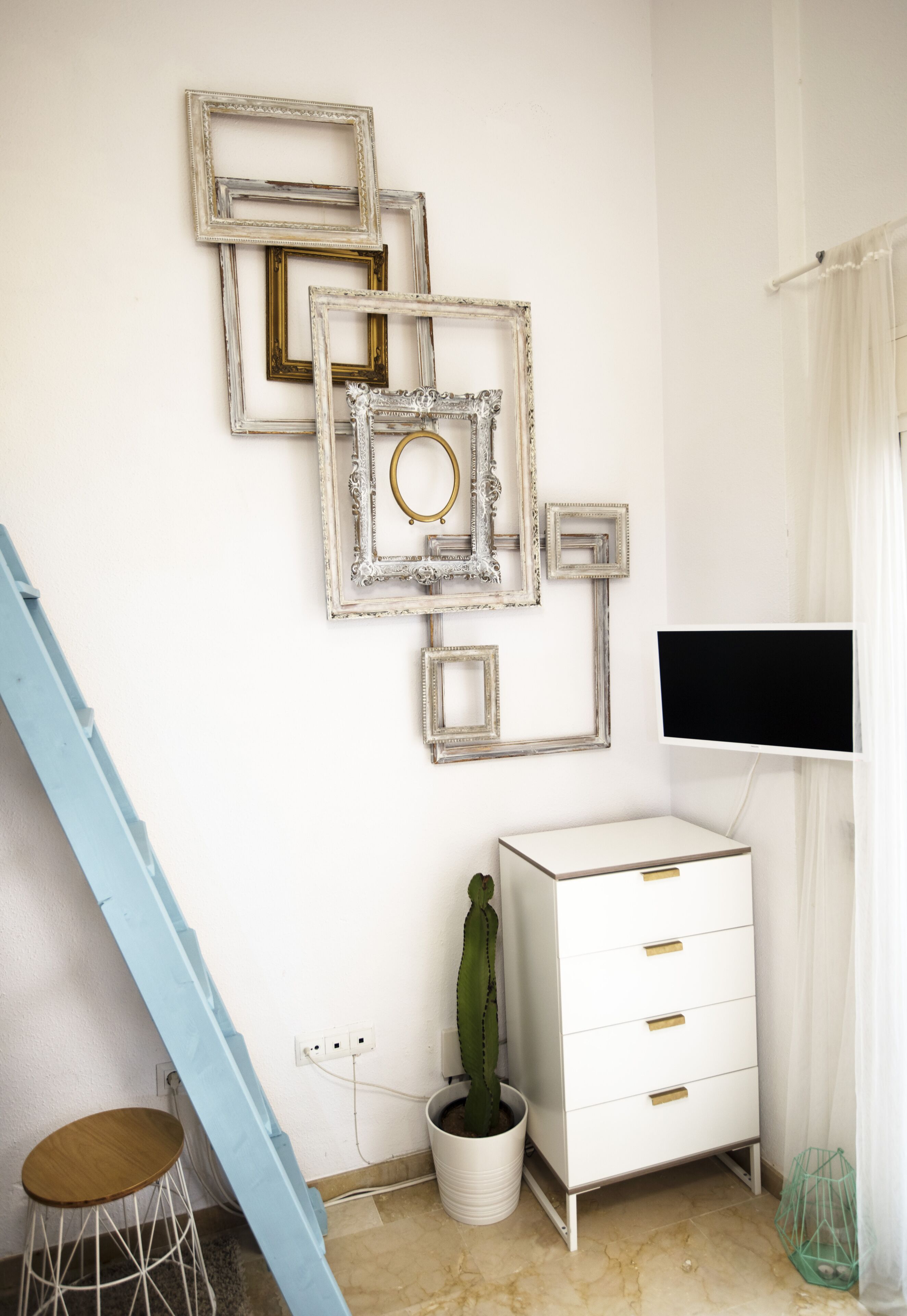 A room with a white wall, a white cabinet with drawers, a cactus in a white pot, a blue chair, and a TV mounted on the wall.