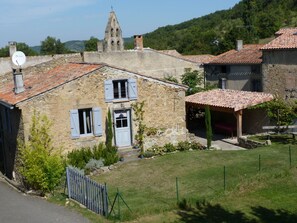 Une maison en pierre avec un toit de tuiles, un petit jardin et un clocher en arrière-plan.