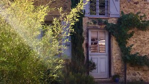 Ein rustikales Steinhaus mit weißer Tür, einer kleinen Veranda und einem Garten mit Bambus und Efeu.