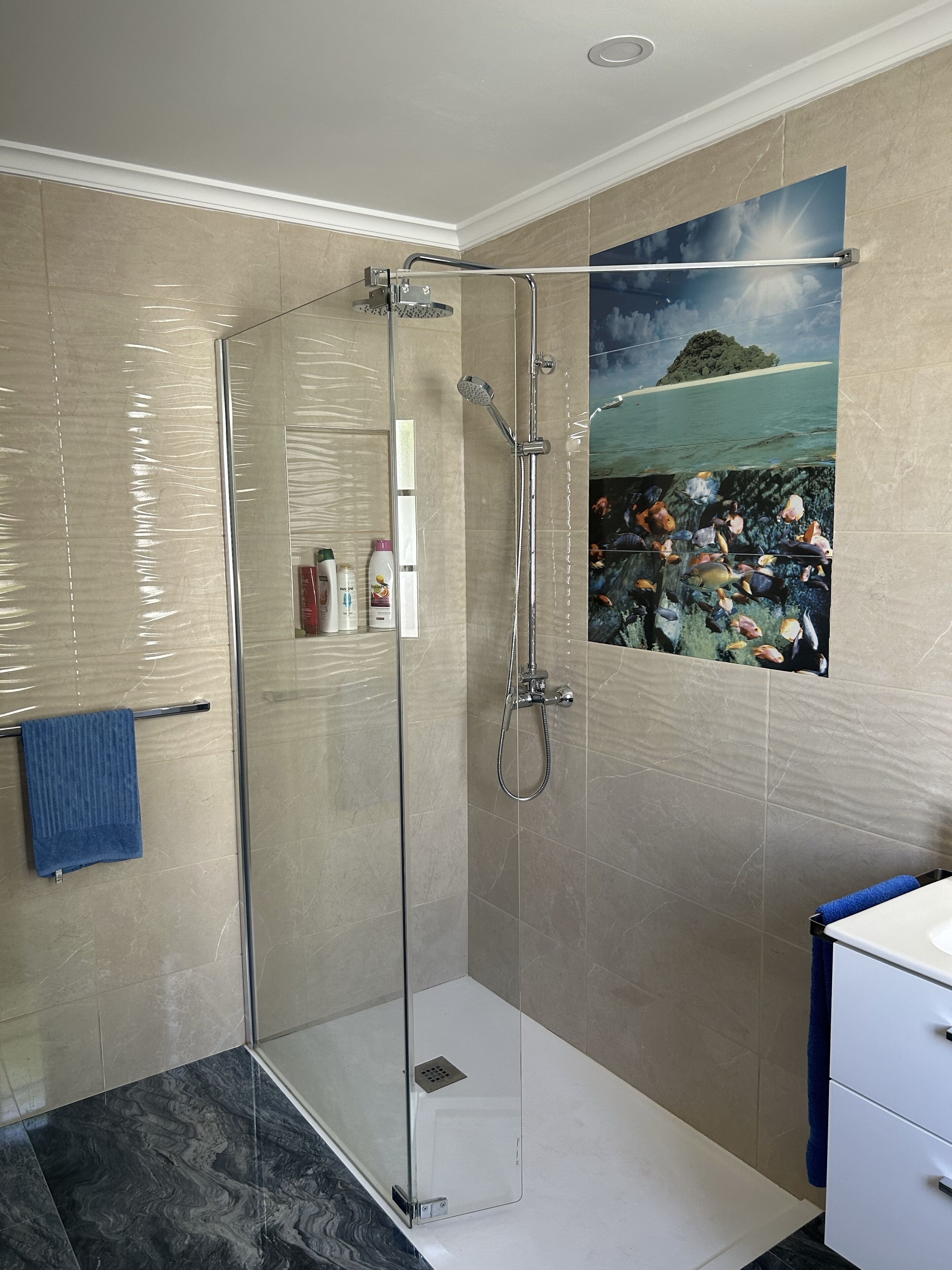 Une salle de bain moderne avec une douche à parois de verre, des carreaux de couleur beige, une serviette bleue et un tableau représentant un paysage tropical.