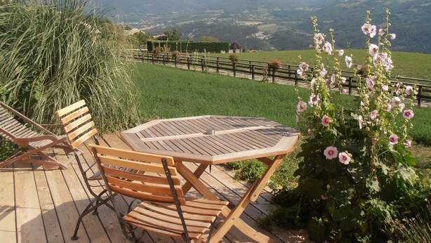 A wooden deck with a table and chairs, overlooking a mountainous landscape with a fence and flowering plants. - Contemporary cottage between Ubaye and Durance overlooking the lake of Serre Ponçon 2 (Le Sauze-Du-Lac)