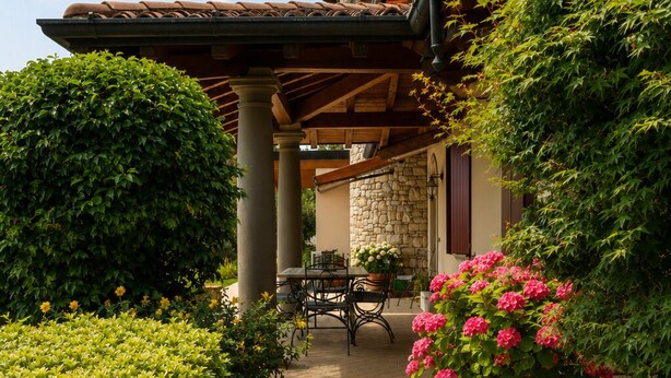 A pathway leading to a covered patio area with a stone wall, a table with chairs, and a potted plant with pink flowers.