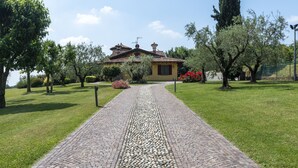 A cobblestone pathway leading to a house with a tiled roof, surrounded by greenery and trees.