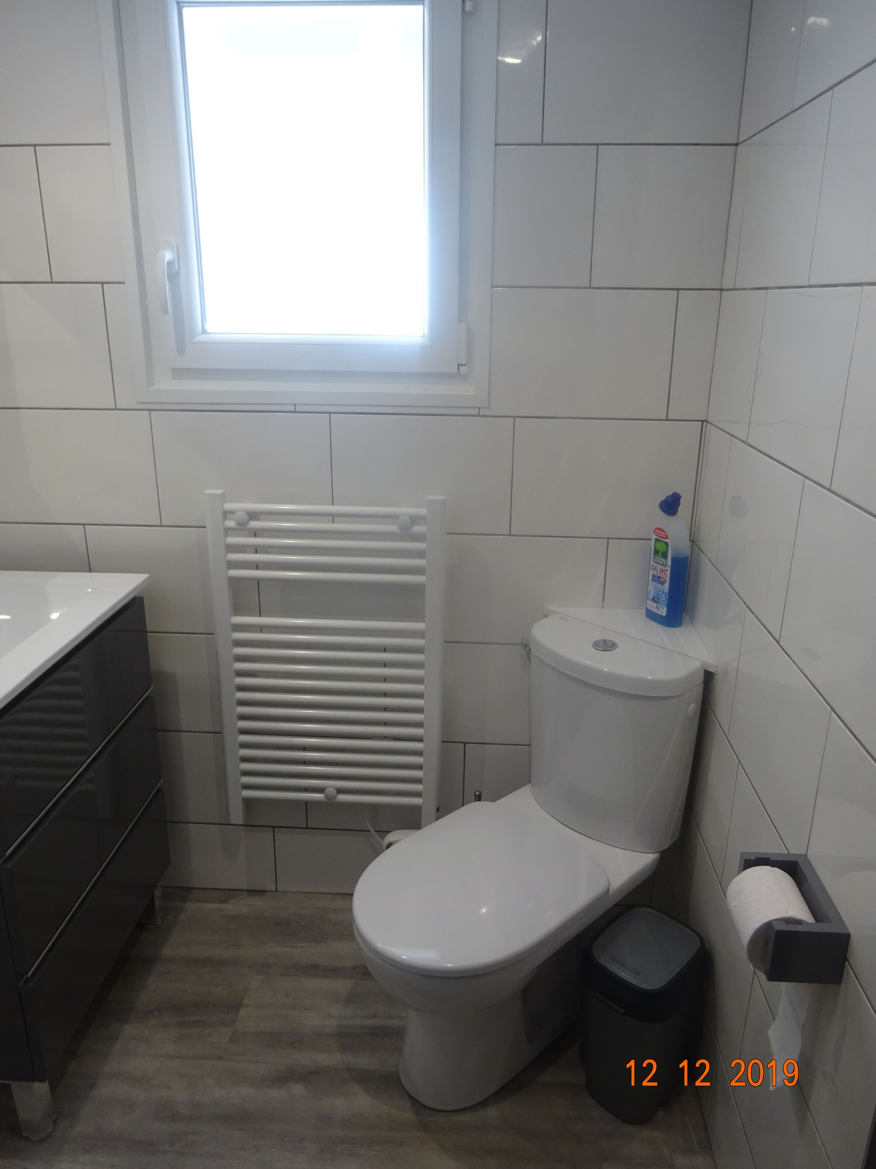 A bathroom with a toilet, a radiator, a window, a sink, and a trash bin.
