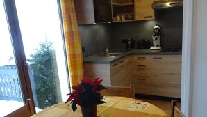 A dining table with a floral tablecloth and a potted plant, a kitchen with wooden cabinets and a coffee machine, and a view through a glass door. - chalet - 4 rooms - 6 persons (La Bresse)