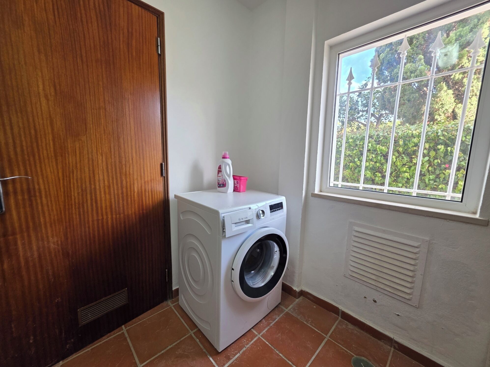 Uma lavanderia compacta com máquina de lavar, uma pequena janela com vista para o verde e uma porta de madeira.