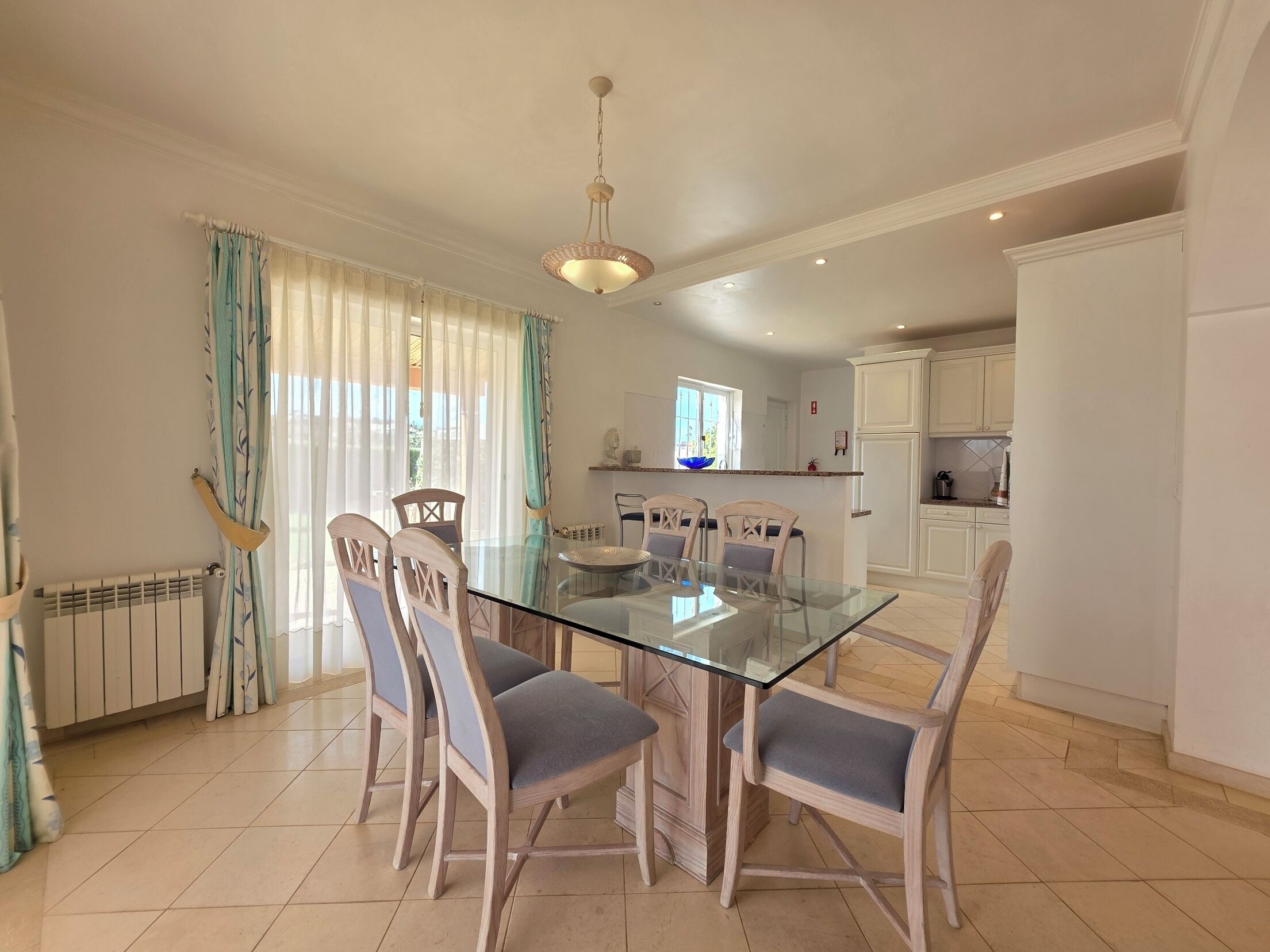 A dining area with a glass table and wooden chairs, a kitchen in the background, and a radiator on the wall.