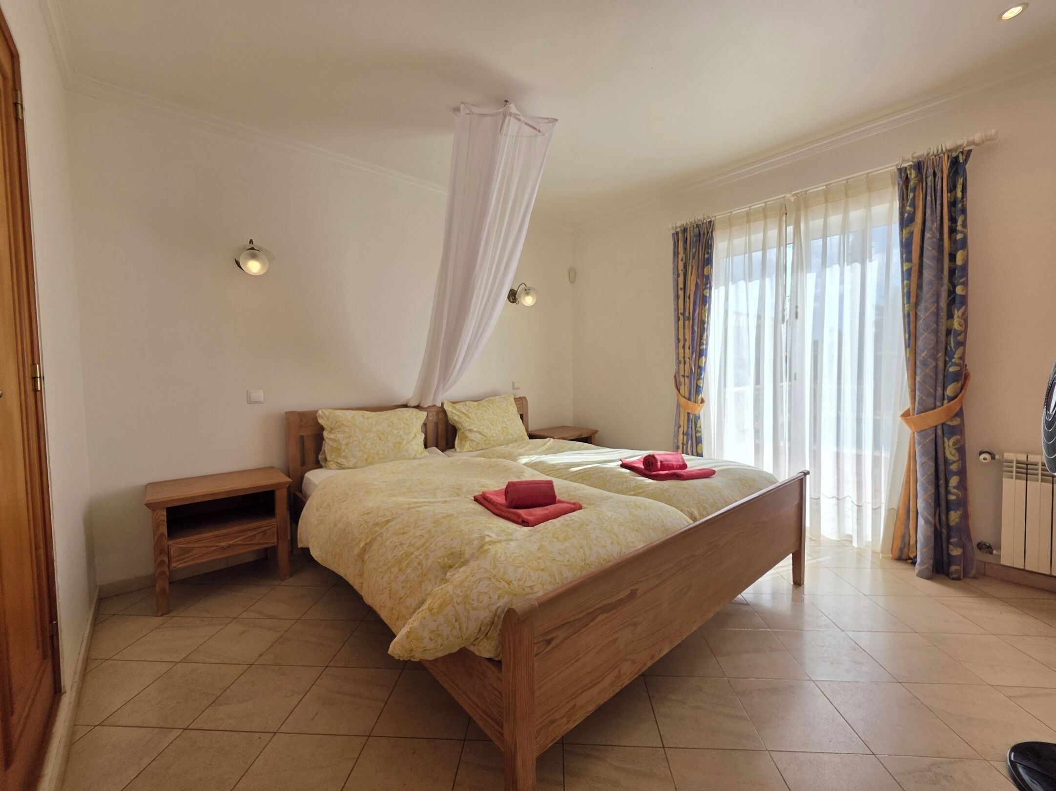 Ein Schlafzimmer mit einem Holzbett, einem Nachttisch, einem Beistelltisch, einem Fenster mit Vorhängen und einer Heizung.