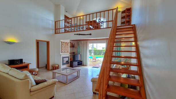 A spacious living room with a high ceiling, a wooden staircase, a fireplace, and a view of the outdoor pool area. - Beautiful villa with private pool & playground near Lagos & the beach (Vale da Lama)