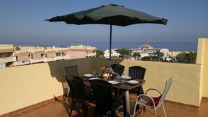 Una terraza con mesa lista para comer, una sombrilla y vistas al mar y edificios.