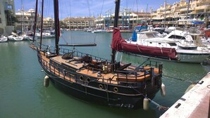 Un gran barco de vela tradicional atracado en una marina, con otros barcos y edificios al fondo.