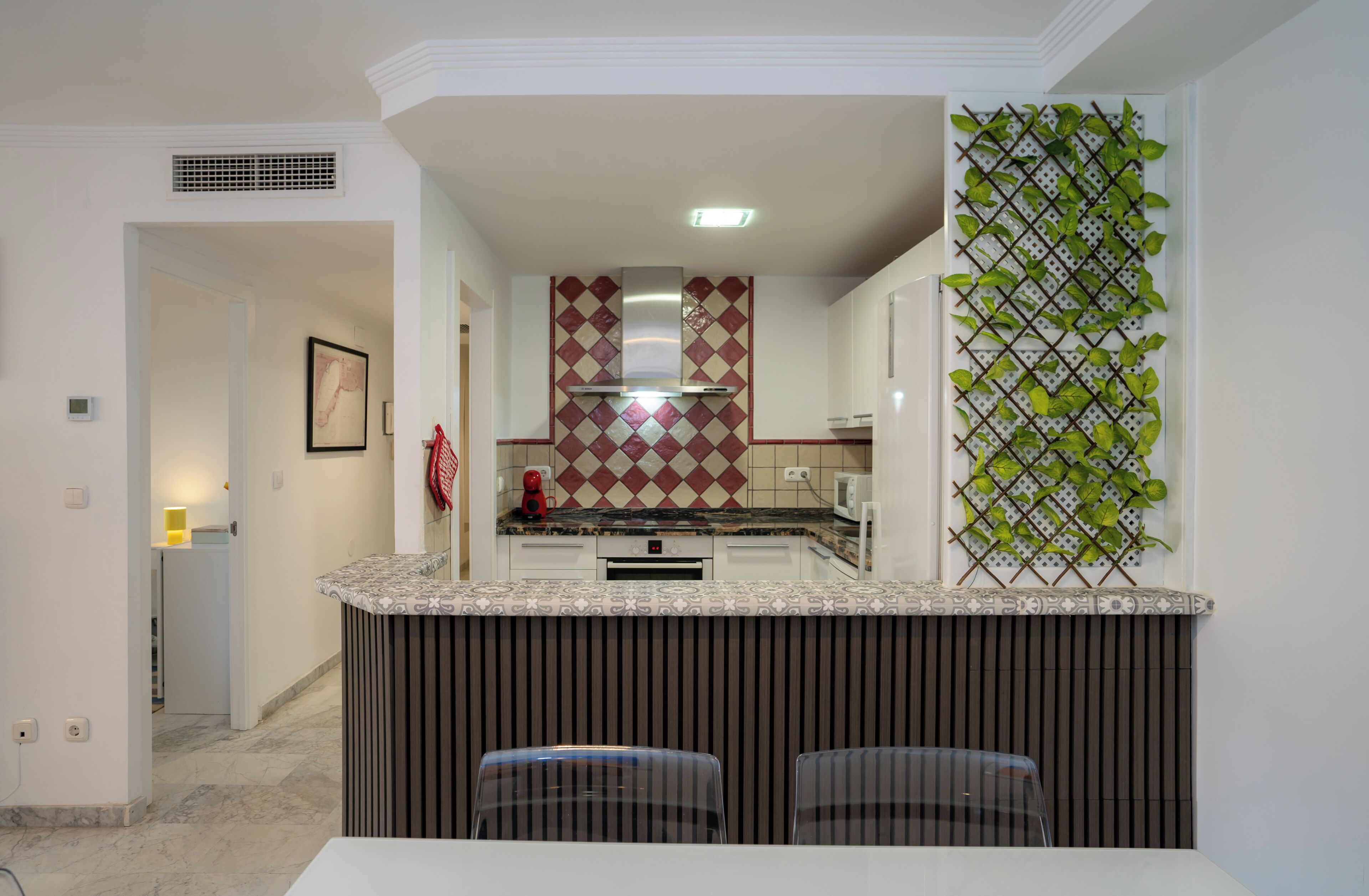 A compact kitchen with a central island, a microwave, and a sink. There is a patterned backsplash and a green plant wall decoration.