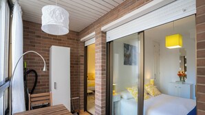 Habitación de hotel con un banco de madera, una cama con ropa de cama amarilla y blanca, un armario blanco y una puerta de vidrio que da a un balcón.