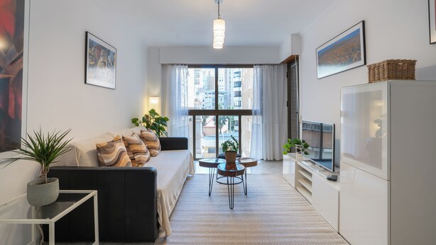 Una sala de estar moderna con un sofá, una mesita, un televisor y un ventanal con cortinas.