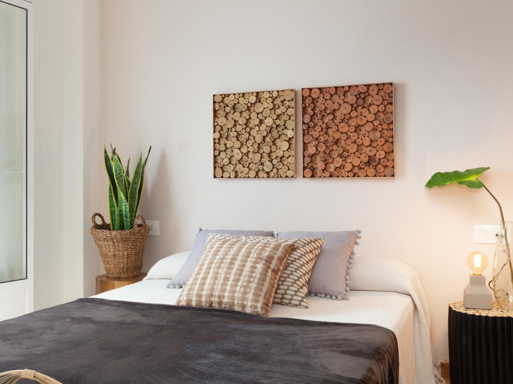 A bedroom with a bed, pillows, a bedside table with a lamp, a potted plant, and two wall-mounted artworks.