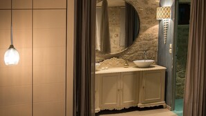 Een slaapkamer met een bed, nachtkastje, lamp en een badkamer met wastafel en spiegel.
