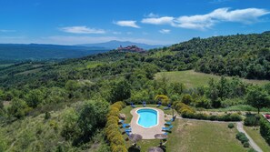 A pool area with lounge chairs and umbrellas, surrounded by lush greenery and a view of a castle on a hill. - Woodland Cottage (A3) with pool - Le Caldanelle (Civitello Paganico, Grosseto)