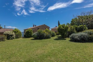 Un paysage rural avec une maison en pierre, une végétation luxuriante et un ciel bleu dégagé.