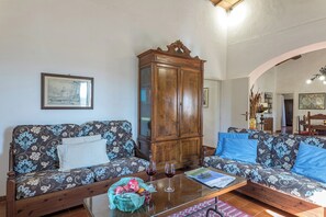 A living room with two patterned sofas, a wooden cabinet, a glass coffee table with a fruit basket and wine glass, and a framed picture on the wall. - Woodland Cottage (A3) with pool - Le Caldanelle (Civitello Paganico, Grosseto)