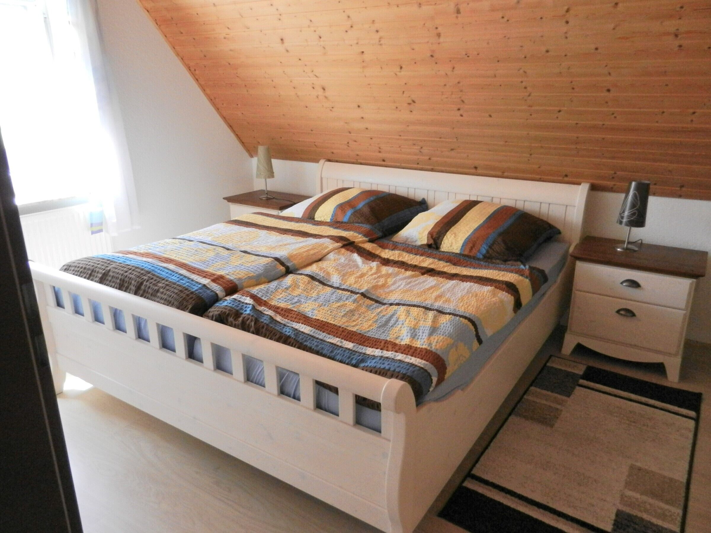 Una camera da letto con soffitto spiovente in legno, un letto bianco con copriletto colorato, un comodino con una lampada e un piccolo mobiletto contenitore.