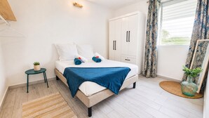 A bedroom with a bed, a wardrobe, a small table with a plant, and a window with curtains. - Beautiful 3-room apartment in the heart of the marina, 3 minutes from the beaches  (Les Trois-Îlets)