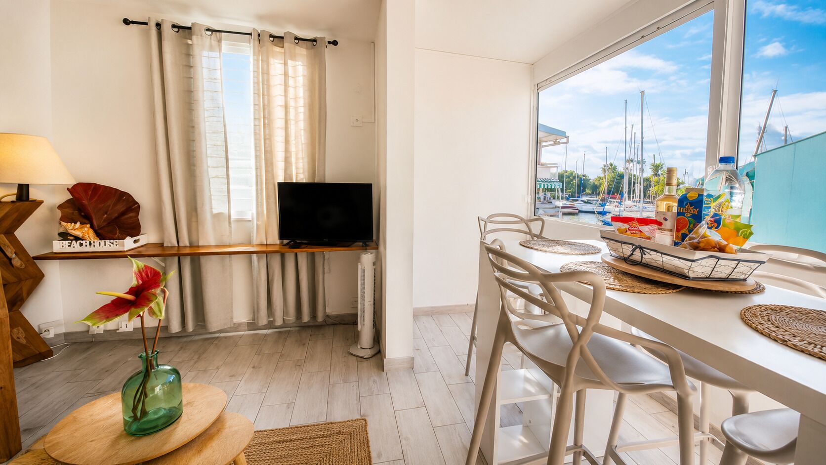 A modern room with a TV, dining table, chairs, a window with a view of boats, and a sign reading 'BEACH HOUSE'.
