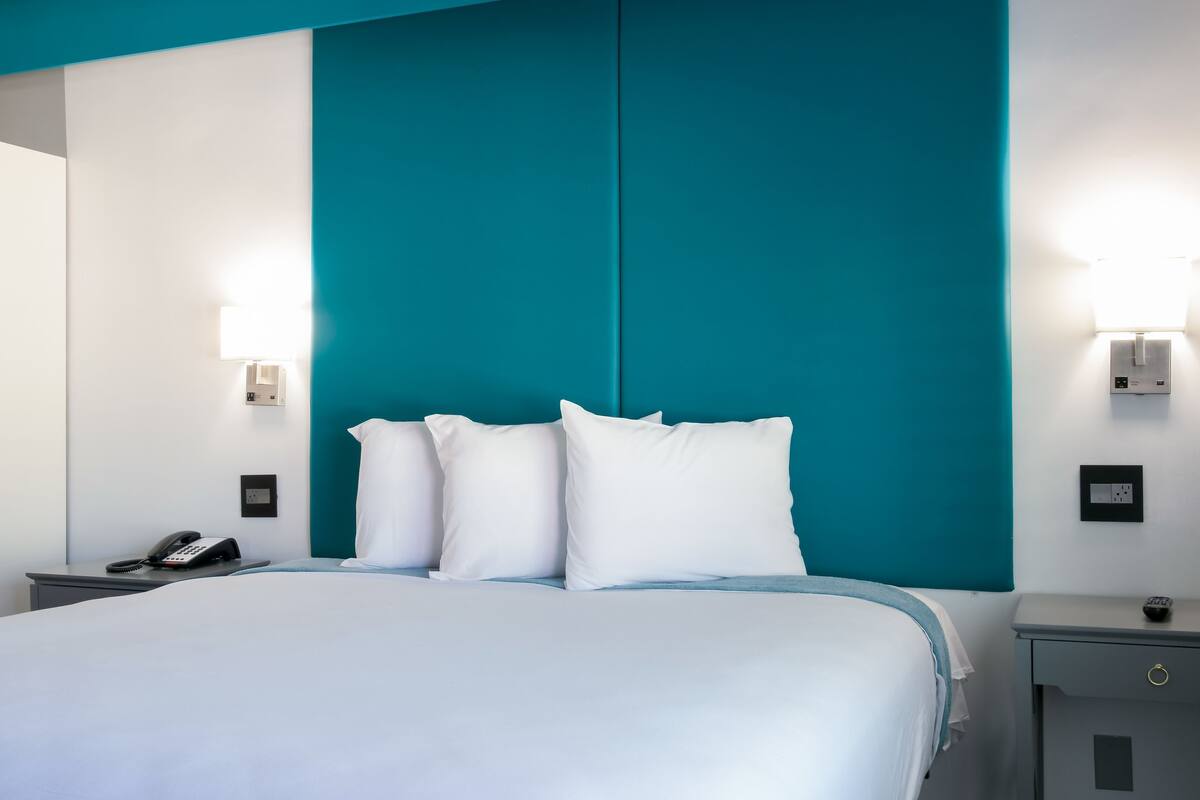 Une chambre d’hôtel avec un lit, du linge de lit blanc, des murs d’une couleur turquoise, des lampes de chevet, un téléphone sur la table de chevet et un petit tiroir.
