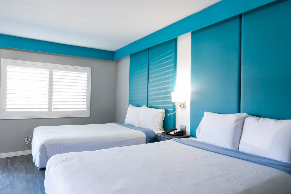 Une chambre d’hôtel avec deux lits, une fenêtre avec des stores, une table de chevet avec un téléphone et un mur d’accent bleu.