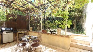 An outdoor patio with a grill, dining table, and chairs, surrounded by a garden and a pergola. - The Ancient Host (Marcoux)