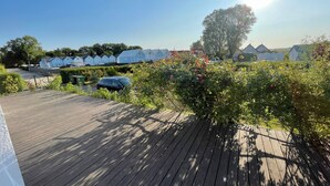 Een houten terras met een geparkeerde auto in de buurt, omringd door groen en een rij huizen op de achtergrond.