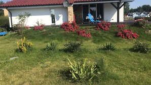 Een huis met een rood pannendak, een overdekte veranda en een tuin met roze bloemen en groene planten.
