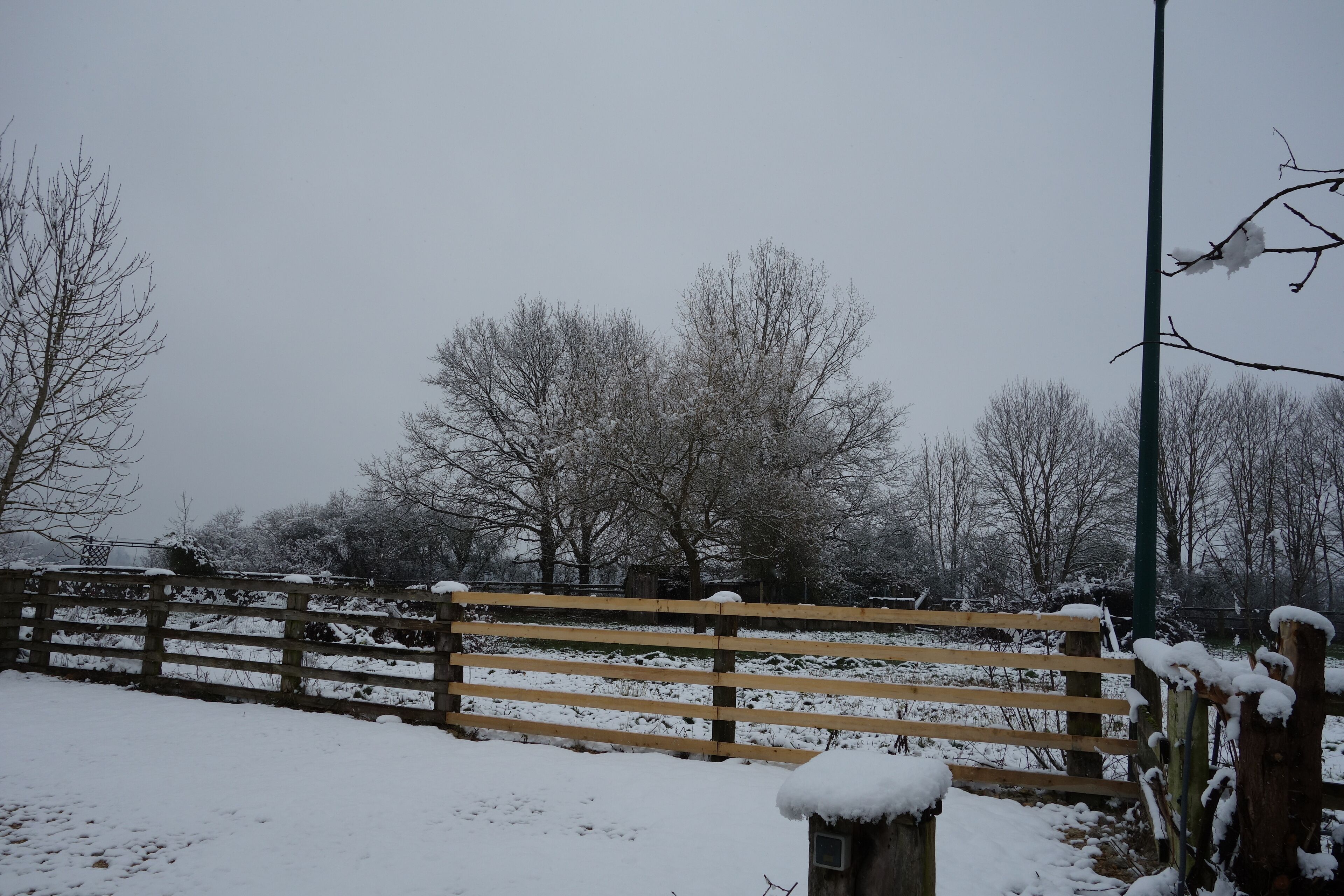 A wooden fence with snow accumulation, a metal post, and bare trees in a snowy landscape.