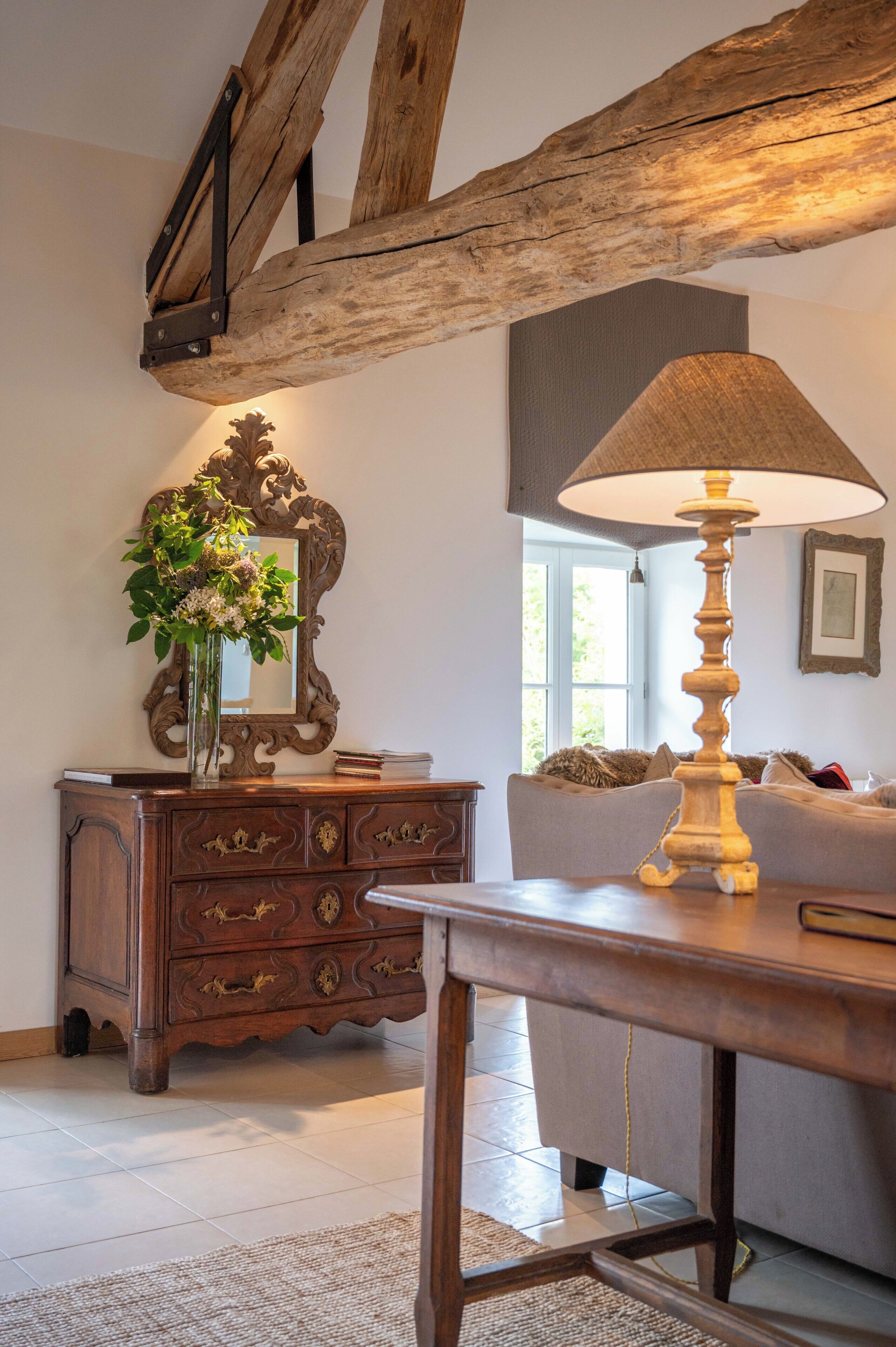 Una habitación con una mesa de madera, una lámpara, un aparador con cajones, un espejo y un sofá.