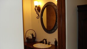 A bathroom with a wooden vanity, a white sink, a mirror with an ornate frame, a wall-mounted light fixture, and a basket with toiletries. - Room 4 Two in Montecito/ Santa Barbara (Montecito)