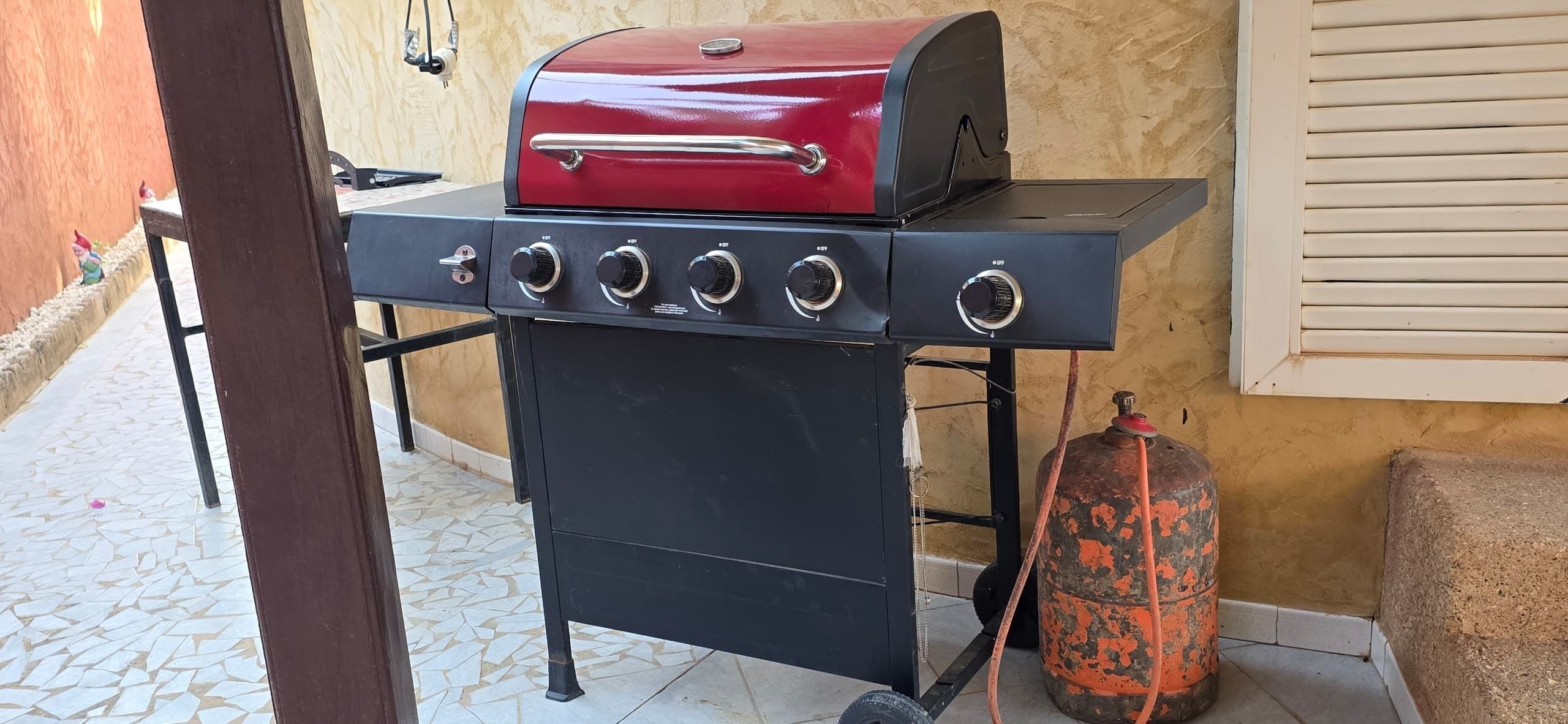 A barbecue grill with a red top and four burners, positioned on a tiled patio with a gas cylinder nearby.