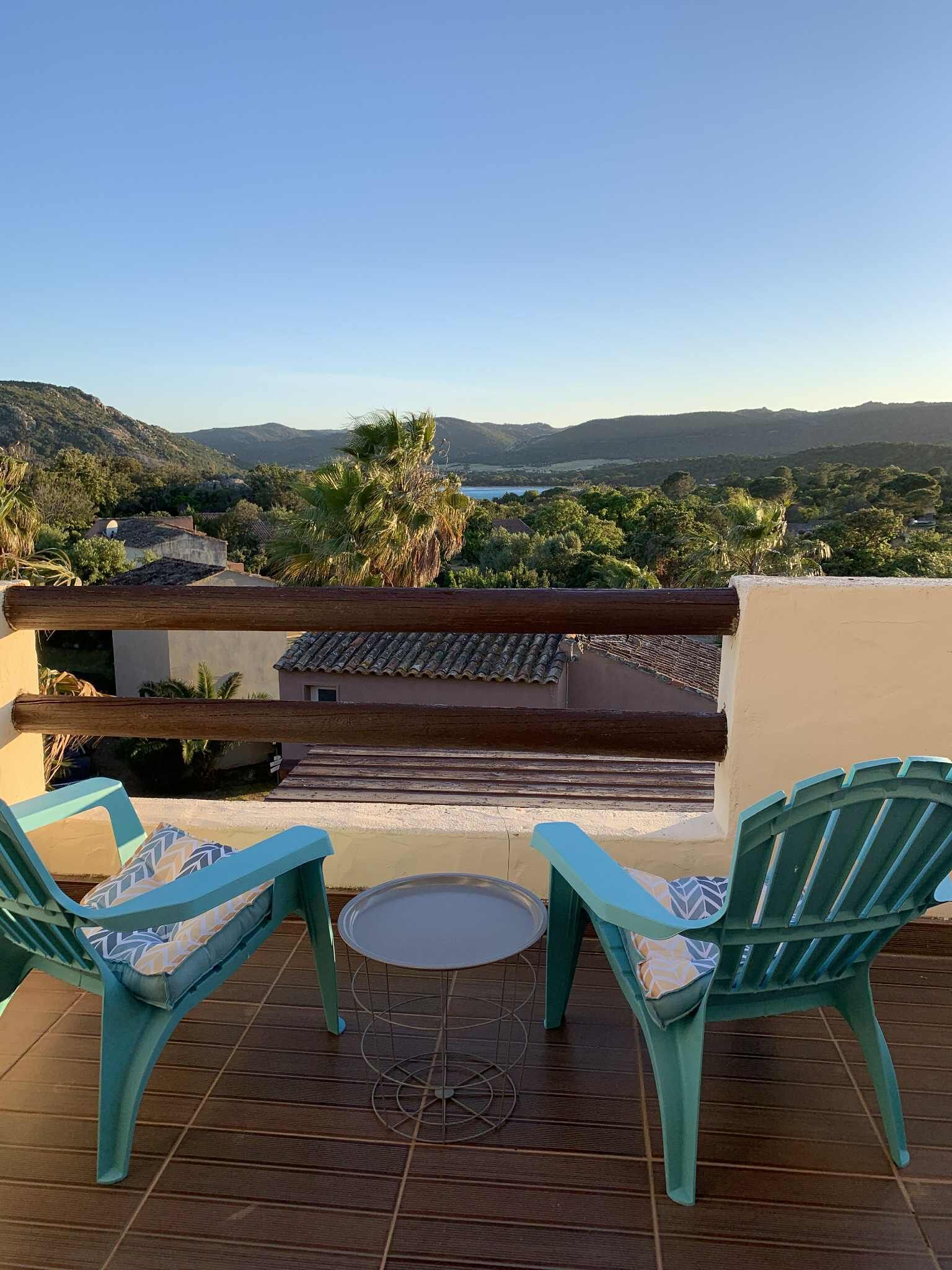 Una terrazza con due poltrone color turchese e un tavolino rotondo, affacciata su un paesaggio pittoresco con colline e uno specchio d'acqua.