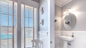 Un baño con un lavamanos blanco de pedestal, un espejo redondo con iluminación y una silla blanca cerca de una ventana con persianas.
