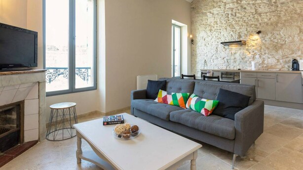 A living room with a grey sofa, a white coffee table, and a fireplace. There is a stone wall and a kitchen in the background. - Nice new 2 bedroom town center apartment (Avignon)