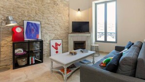 A living room with a stone wall, a fireplace, a flat-screen TV, a sofa with colorful cushions, and a coffee table with books and a bowl of snacks. - Nice new 2 bedroom town center apartment (Avignon)