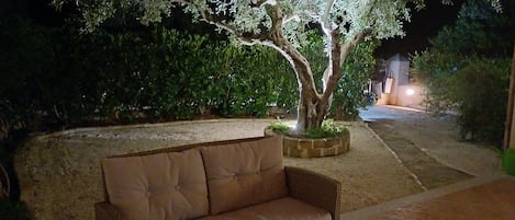 Ein Korbsessel mit Kissen, ein Baum in einem Blumentopf und eine gepflasterte Fläche mit Mauerwerk und Grünpflanzen im Hintergrund.